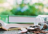 Pile of money coins in the glass jar with books on blurred natural green background and added colour filter for financial and education concept