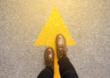 Feet and arrows on road background in starting line beginning idea. Top view. Woman in leather ankle boots on pathway with yellow direction arrow symbol. Moving forward, new start and success concept.