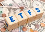 Wooden cubes with blue letters forming the word ETFS (Exchange Traded Funds) on dollar bills on a table. Investment concept photo.