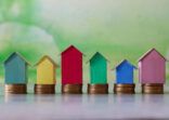 Close-up image of row multi-coloured cardboard houses, colourful beach huts stood on piles of coins, focus on foreground, mottled green background, family finances, real estate and holiday savings concept