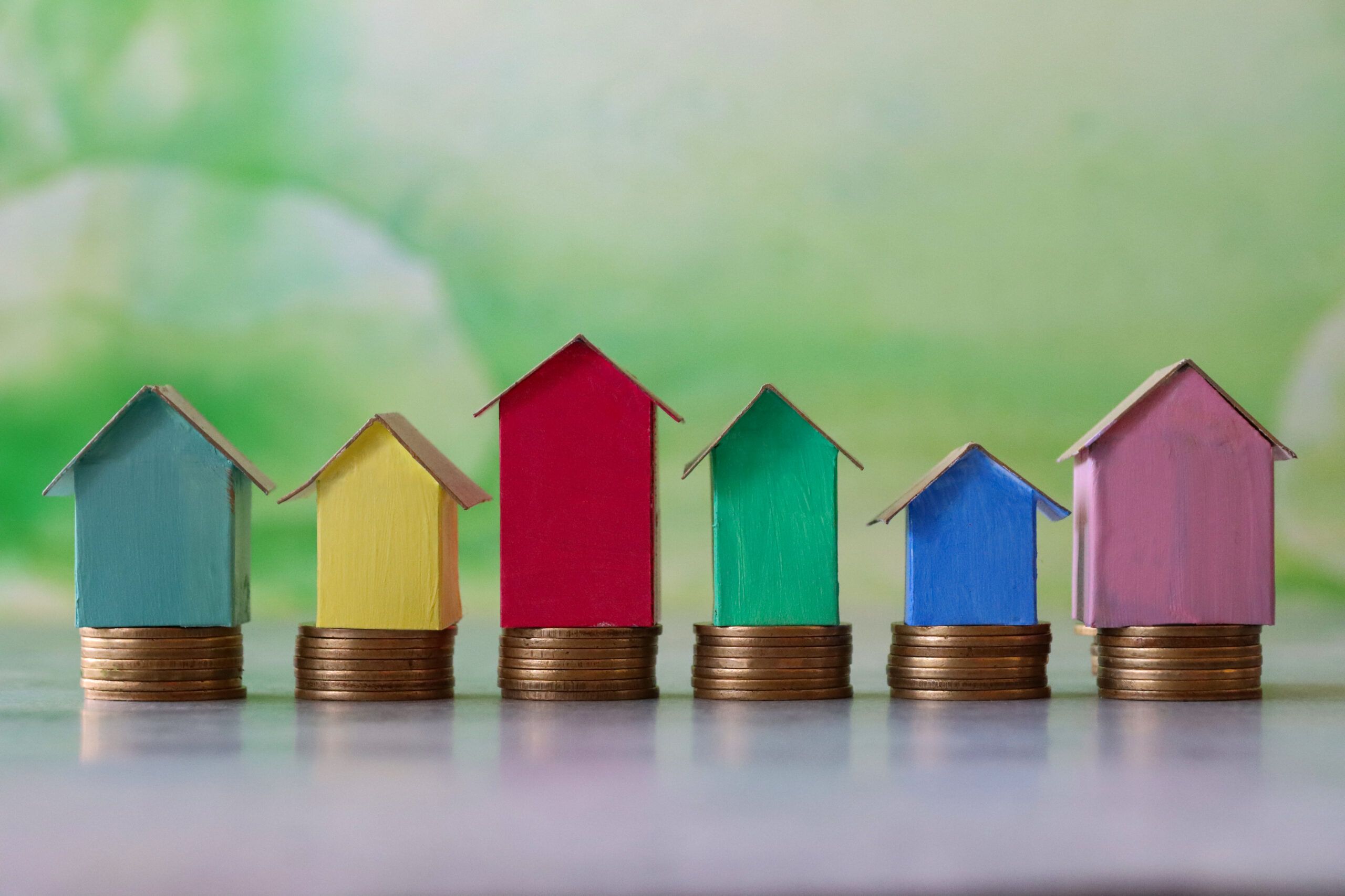 Close-up image of row multi-coloured cardboard houses, colourful beach huts stood on piles of coins, focus on foreground, mottled green background, family finances, real estate and holiday savings concept