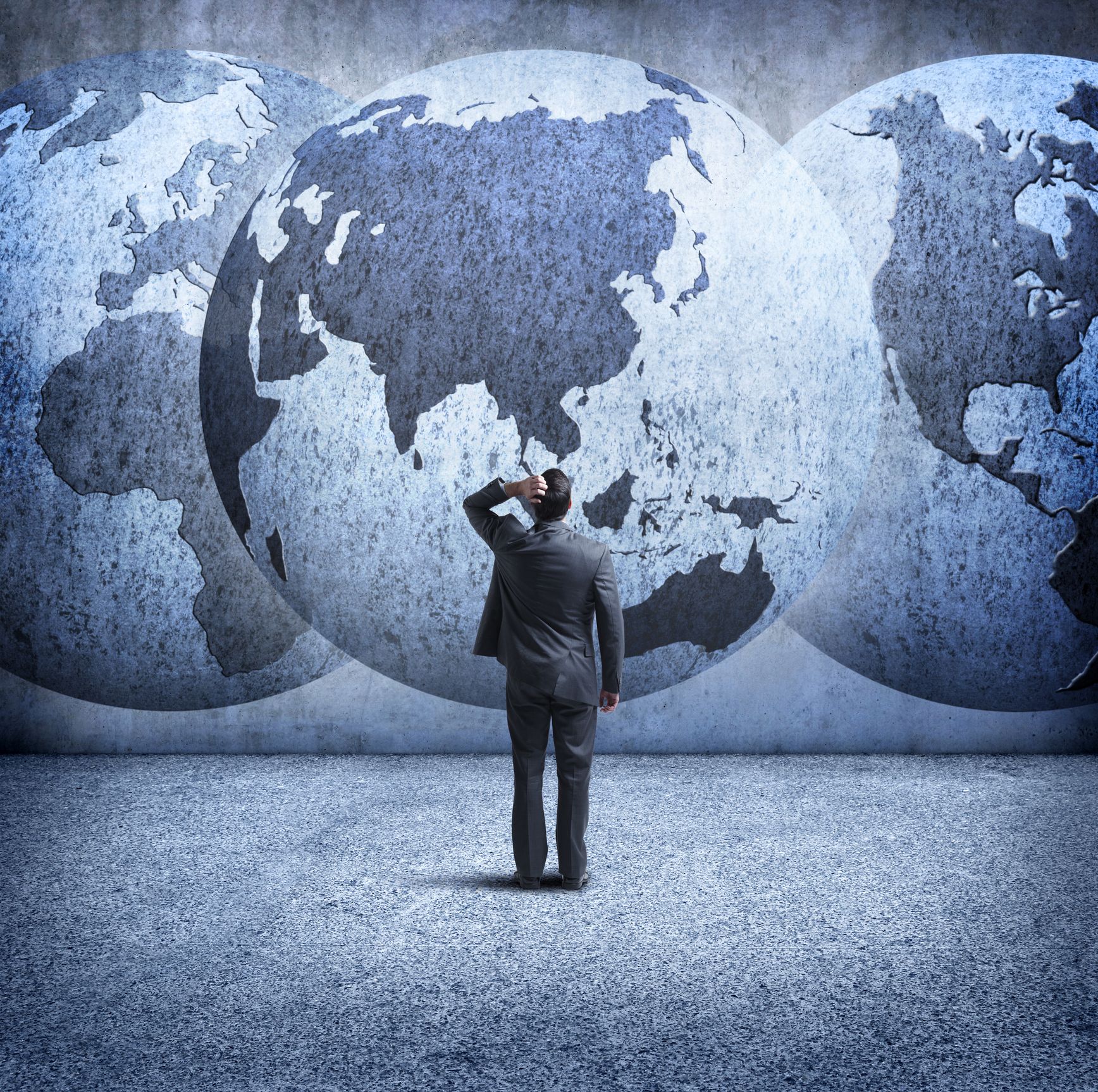 Businessman Stands And Looks Up At Three Interlocking Globes On Wall